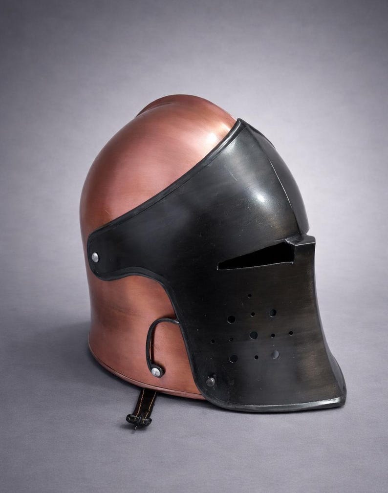 May include: A copper-colored medieval helmet with a black visor and face guard. The helmet has a rounded top and a riveted construction. The visor features small eye slits and ventilation holes. A leather strap with a buckle is attached to the helmet.