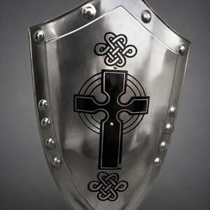 Pode incluir: Um escudo polido, prateado, com uma base pontiaguda e um topo arredondado. O escudo apresenta um desenho de cruz celta preta no centro, com nós decorativos acima e abaixo. Rebites prateados alinham as bordas.