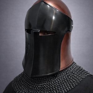 May include: A medieval helmet with a copper-colored top and a black face shield. The helmet has a visor with eye slits and a chin guard. A chainmail collar is visible below the helmet.