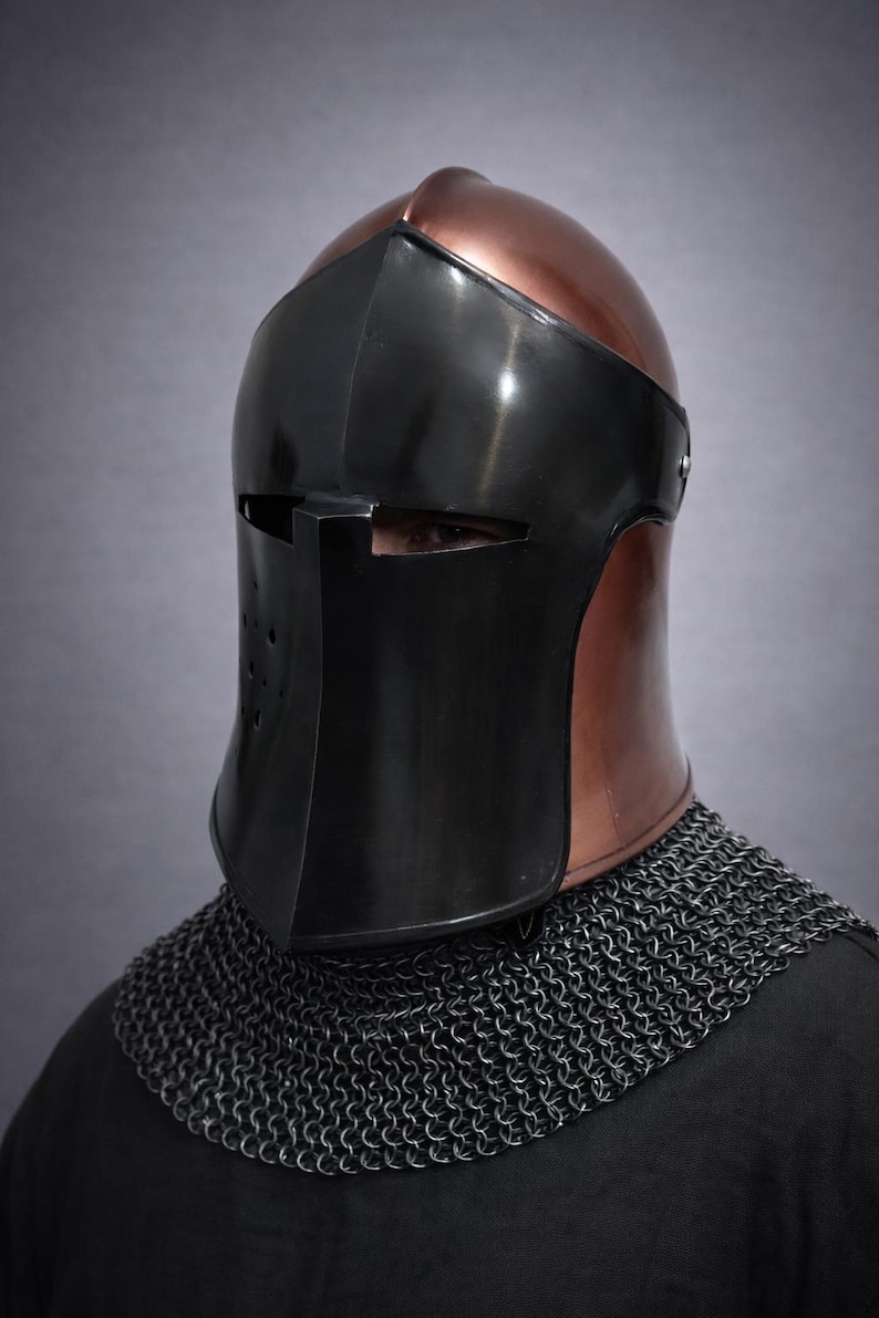 May include: A medieval helmet with a copper-colored top and a black face shield. The helmet has a visor with eye slits and a chin guard. A chainmail collar is visible below the helmet.