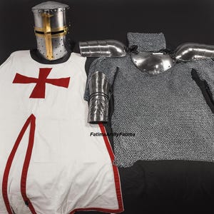 May include: Medieval knight's armor set. The set includes a silver helmet with a gold cross, a white tunic with a red cross, chainmail, and metal shoulder and arm guards. The armor is displayed on a black surface.