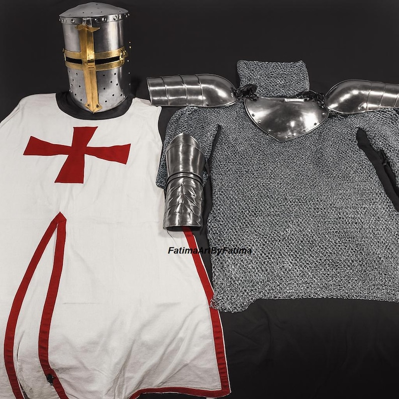 May include: Medieval knight's armor set. The set includes a silver helmet with a gold cross, a white tunic with a red cross, chainmail, and metal shoulder and arm guards. The armor is displayed on a black surface.