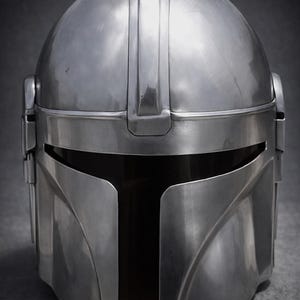 Może przedstawiać: Srebrny hełm Mandalorian z gładkim, metalicznym wykończeniem. Hełm posiada wizjer w kształcie litery T i charakterystyczny, zakrzywiony kształt. Powierzchnia odbija światło, podkreślając jego kontury i detale.