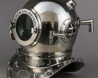 Puede incluir: Un casco de buceo de metal plateado pulido con una gran ventana frontal circular que tiene el texto ANCHOR ENGINEERING grabado en el borde, ventanas laterales, una ventilación superior y una placa base ancha con múltiples pernos.