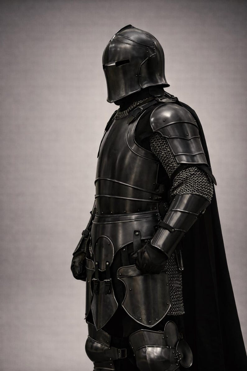 May include: A full suit of medieval armor in dark metal, including a helmet, chest plate, and leg guards. The armor features chainmail and a black cape. The image is a side view of the armor.