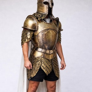 Puede incluir: Una armadura completa de color oro antiguo, que incluye un casco, protectores de hombros, peto y armadura inferior en forma de falda. La armadura tiene detalles intrincados y una capa. La persona lleva pantalones cortos negros.