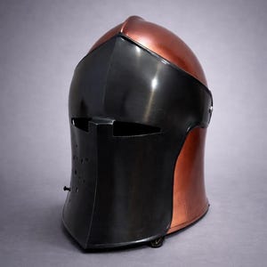 May include: A medieval-style helmet with a copper-colored top and a black face shield. The helmet has a rounded shape with a visor and a series of small holes on the lower front. The design is reminiscent of historical armor.