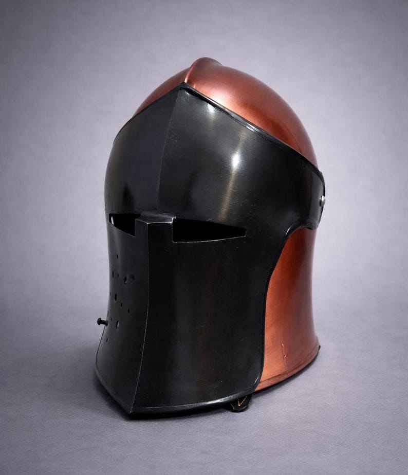 May include: A medieval-style helmet with a copper-colored top and a black face shield. The helmet has a rounded shape with a visor and a series of small holes on the lower front. The design is reminiscent of historical armor.