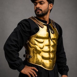 May include: A gold-coloured metallic breastplate featuring a moulded anatomical muscle design on the front. The armour piece includes dark leather shoulder straps with metal rivets and adjustable side buckles, secured with a black cord at the neckline.