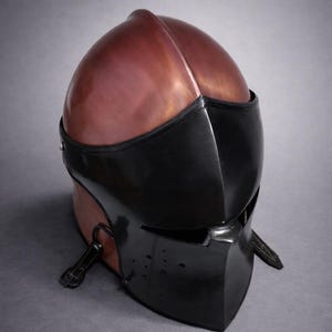 May include: A medieval-style helmet with a copper-colored top and a black face guard. The helmet has a rounded shape with a central ridge and a visor with eye slits. Leather straps are attached to the sides.