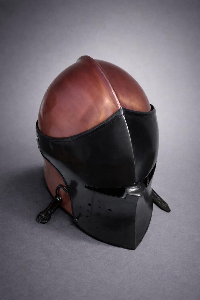 May include: A medieval-style helmet with a copper-colored top and a black face guard. The helmet has a rounded shape with a central ridge and a visor with eye slits. Leather straps are attached to the sides.