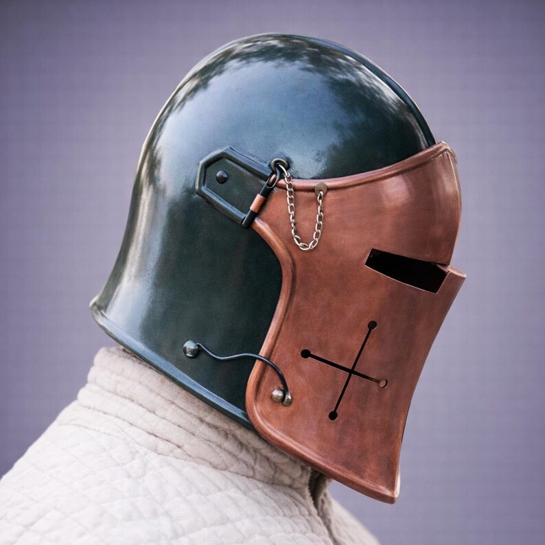 Puede incluir: Un casco de estilo medieval con una parte superior verde oscuro y una placa frontal de color cobre. La placa frontal presenta un dise&ntilde;o de cruz y ranuras para los ojos. Una peque&ntilde;a cadena conecta la placa frontal al casco. El casco se lleva sobre una prenda blanca.
