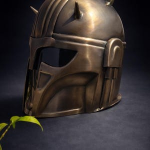 K&ouml;nnte beinhalten: Ein bronzefarbener Helm mit Visier und zwei spitzen H&ouml;rnern oben. Der Helm hat ein detailliertes Design mit Rippen und kantigen Merkmalen, was auf ein futuristisches oder Fantasy-Thema hindeutet. Der Helm wird vor einem dunklen Hintergrund pr&auml;sentiert.