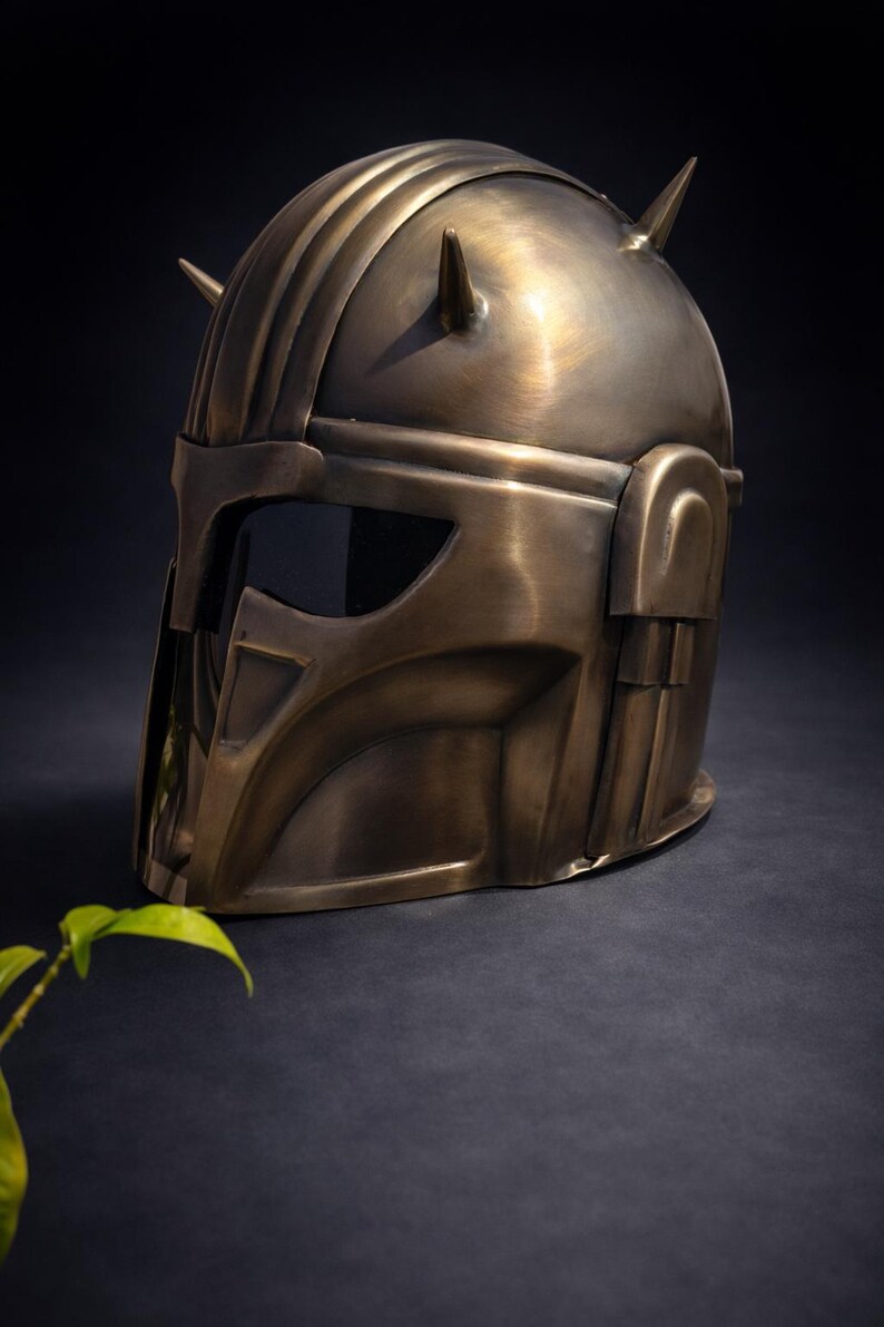 K&ouml;nnte beinhalten: Ein bronzefarbener Helm mit Visier und zwei spitzen H&ouml;rnern oben. Der Helm hat ein detailliertes Design mit Rippen und kantigen Merkmalen, was auf ein futuristisches oder Fantasy-Thema hindeutet. Der Helm wird vor einem dunklen Hintergrund pr&auml;sentiert.