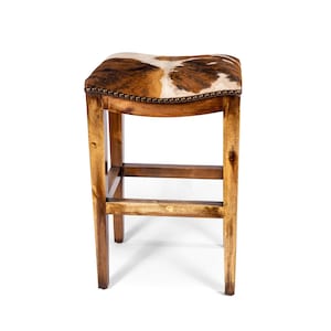 Pode incluir: Banqueta de bar de madeira com assento de pele de vaca. O assento &eacute; marrom, branco e bege. A estrutura de madeira &eacute; de cor marrom claro. O assento &eacute; adornado com pequenos tachas redondas de cor bronze. A banqueta tem um apoio para os p&eacute;s.