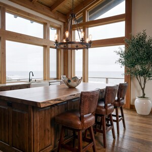May include: A rustic kitchen island with a live edge wooden countertop and dark wood cabinetry. Three leather bar stools with studded backs are positioned at the island. A decorative chandelier hangs above. A large window overlooks the ocean.
