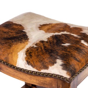 Pode incluir: Banqueta quadrada de madeira com assento de pele de vaca. O assento apresenta um padr&atilde;o castanho, branco e bege. A estrutura de madeira &eacute; castanha m&eacute;dia e o assento &eacute; adornado com pequenos pregos redondos de cor bronze.