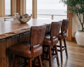 May include: Three brown leather bar stools with a crocodile pattern on the backs are positioned at a wooden kitchen island. The stools have dark wooden legs and are set against a backdrop of a coastal view.