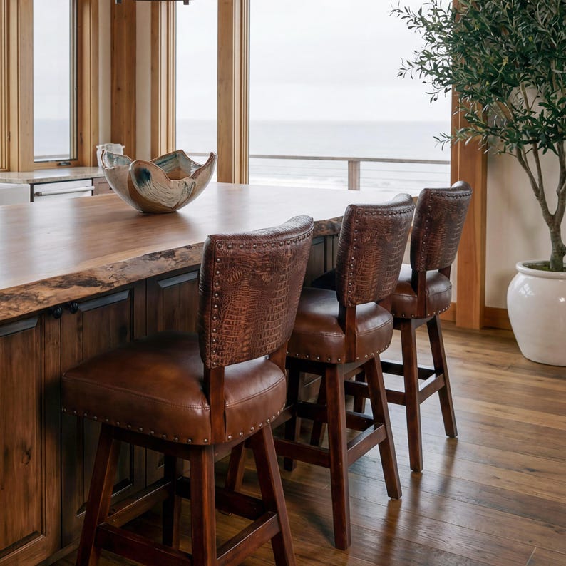 May include: Three brown leather bar stools with a crocodile pattern on the backs are positioned at a wooden kitchen island. The stools have dark wooden legs and are set against a backdrop of a coastal view.