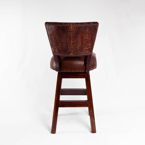 May include: A brown bar stool with a dark wood frame and a leather seat and back. The back of the stool has a textured, crocodile-like pattern. The stool has a footrest and is set against a white background.