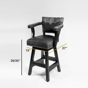 May include: A dark gray swivel bar stool with a faux leather seat and backrest. The backrest features a decorative embossed pattern. The stool has armrests and a footrest. The seat is 19 inches wide, and the overall height is 26/30 inches. The stool is 27 inches wide.