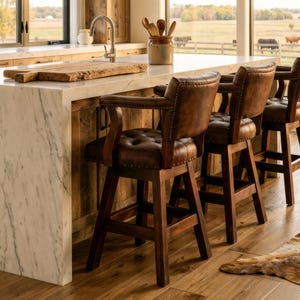 May include: Three brown leather bar stools with armrests are positioned at a kitchen island. The stools have button-tufted backs and dark wooden frames. The kitchen features a marble countertop and a wooden backsplash, with a view of a rural landscape.