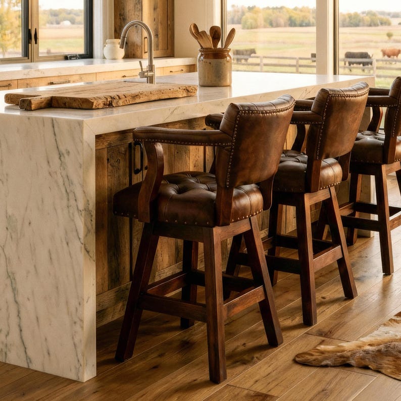 May include: Three brown leather bar stools with armrests are positioned at a kitchen island. The stools have button-tufted backs and dark wooden frames. The kitchen features a marble countertop and a wooden backsplash, with a view of a rural landscape.