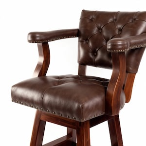 May include: A brown leather bar stool with a tufted back and seat, featuring wooden armrests and a sturdy wooden base. The chair has a classic design with decorative nailhead trim along the edges, suitable for a home bar or kitchen.