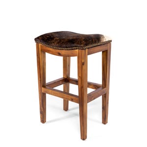 Pode incluir: Banqueta de bar de madeira com assento de pele de vaca. O assento &eacute; marrom e branco com um design curvo. A estrutura &eacute; feita de madeira marrom clara. A banqueta tem quatro pernas e um apoio para os p&eacute;s.
