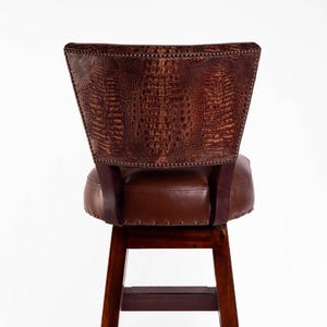 May include: A brown leather bar stool with a crocodile-textured backrest. The seat and back are a darker brown, with a wooden frame and legs. The backrest is studded with small metal tacks.
