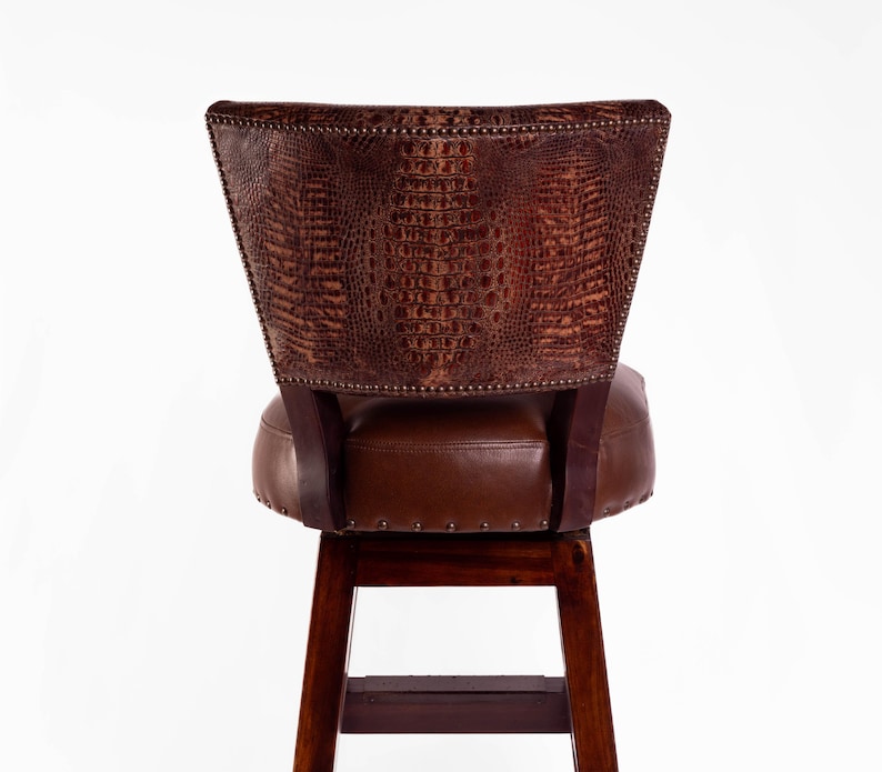 May include: A brown leather bar stool with a crocodile-textured backrest. The seat and back are a darker brown, with a wooden frame and legs. The backrest is studded with small metal tacks.
