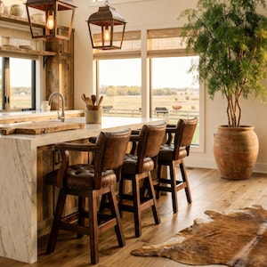 May include: A rustic kitchen with a marble countertop and wooden cabinets. Three brown leather bar stools with armrests are positioned at the counter. Two hanging lanterns with Edison bulbs illuminate the space. A large potted plant and a cowhide rug add to the decor.