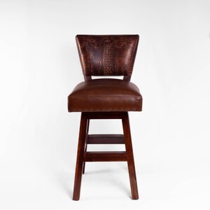 May include: A brown leather bar stool with a dark wood frame. The backrest features a textured, embossed pattern, and the seat is padded. The stool has a footrest and is designed for comfort and style.