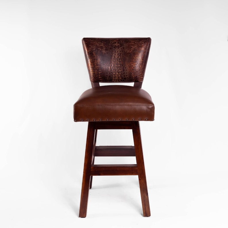 May include: A brown leather bar stool with a dark wood frame. The backrest features a textured, embossed pattern, and the seat is padded. The stool has a footrest and is designed for comfort and style.
