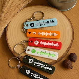 Pode incluir: Vários chaveiros coloridos, cada um com um design de código Spotify. Os chaveiros retangulares têm bordas arredondadas e um chaveiro de metal. As cores incluem azul claro, laranja, vermelho, branco e preto. Os chaveiros estão dispostos em uma superfície de madeira.