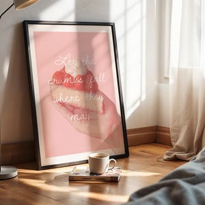 Könnte beinhalten: Gerahmter Kunstdruck mit rosa Hintergrund und dem Schriftzug "Let the crumbs fall where they may". Der Druck zeigt eine Aquarellillustration einer Hand, die ein Stück Kuchen hält. Eine Kaffeetasse und ein Buch liegen auf dem Boden.