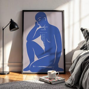 Könnte beinhalten: Ein gerahmter Kunstdruck mit einer blauen Figur in sitzender Pose. Das Kunstwerk trägt den Text "Lapis Lazuli" und weiteren Text in einer vertikalen Spalte. Eine schwarze Lampe, eine Kaffeetasse und ein Buch befinden sich im Vordergrund.