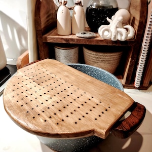 Op de afbeelding: Een houten vergiet met een geperforeerd oppervlak staat op een gespikkelde blauw-grijze pot. De vergiet heeft een donkerhouten handvat. Andere keukenartikelen zijn zichtbaar op de achtergrond, waaronder een houten plank met decoratieve flessen en een klein octopusbeeldje.