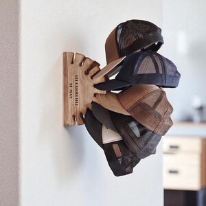 May include: A wooden hat rack mounted on a white wall, holding several baseball caps. The caps are various colors, including brown, navy, and black, with mesh backs. The rack has a unique design with slots for each hat and the text "LES CAQUETTES FT MAX".