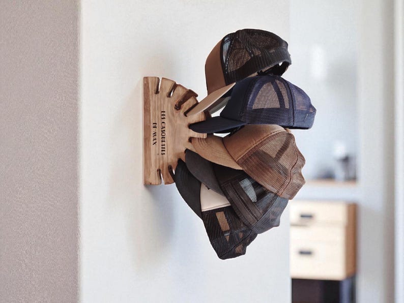 May include: A wooden hat rack mounted on a white wall, holding several baseball caps. The caps are various colors, including brown, navy, and black, with mesh backs. The rack has a unique design with slots for each hat and the text "LES CAQUETTES FT MAX".