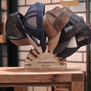 May include: A wooden hat display stand with several trucker caps. The caps are in shades of black, navy, and brown, featuring mesh backs and curved peaks. The stand is engraved with the text "LES CASQUETTES DE MAX".