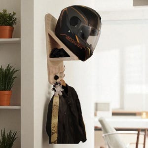 Può includere: Un portacasco e attaccapanni a parete in legno. Un casco da moto nero e oro poggia su un ripiano. Sotto, una giacca nera è appesa a un gancio, insieme a un piccolo peluche. Il portacasco è montato su una parete bianca.