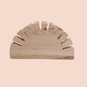 May include: A light-colored wooden wine glass holder, semi-circular in shape. The holder has multiple slots cut into the top to hold wine glasses. The wood grain is visible, and the piece is set against a light pink background.