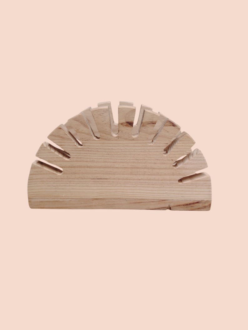 May include: A light-colored wooden wine glass holder, semi-circular in shape. The holder has multiple slots cut into the top to hold wine glasses. The wood grain is visible, and the piece is set against a light pink background.