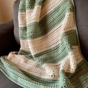May include: A handmade crochet blanket in soft green and cream colors. The blanket features a striped pattern with textured stitches and a decorative border. The blanket is draped over a gray sofa, showcasing its cozy and inviting design.