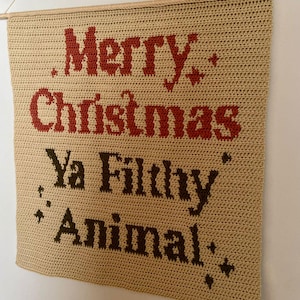May include: A beige crocheted wall hanging with the words "Merry Christmas Ya Filthy Animal" in red and brown. The text is centered on the rectangular piece, which is suspended from a wooden dowel.