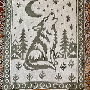 Puede incluir: Una manta de ganchillo rectangular en tonos verde salvia y crema. El diseño muestra un lobo aullando a una luna creciente rodeado de estrellas, pinos y montañas. El borde tiene un patrón geométrico y flecos en los extremos.