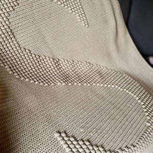 Könnte beinhalten: Eine beige Strickdecke mit strukturiertem Muster. Die Decke hat ein sich wiederholendes geometrisches Design mit erhabenen, knötchenartigen Details. Die Gesamtästhetik ist gemütlich und einladend, passend für die Inneneinrichtung.