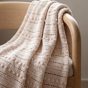 Könnte beinhalten: Eine hellbeige Häkeldecke mit einem strukturierten Muster aus horizontalen Reihen, das zwischen festen Maschen, durchbrochenen Spitzenabschnitten und kleinen erhabenen Noppenclustern im gesamten Stoff wechselt, drapiert über einem Holzstuhl.