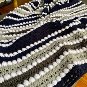 May include: A handmade crochet blanket with a striped pattern. The blanket features alternating rows of navy blue, gray, brown, and white. White bobbles are evenly spaced along the rows, adding texture and visual interest to the cozy, soft blanket.